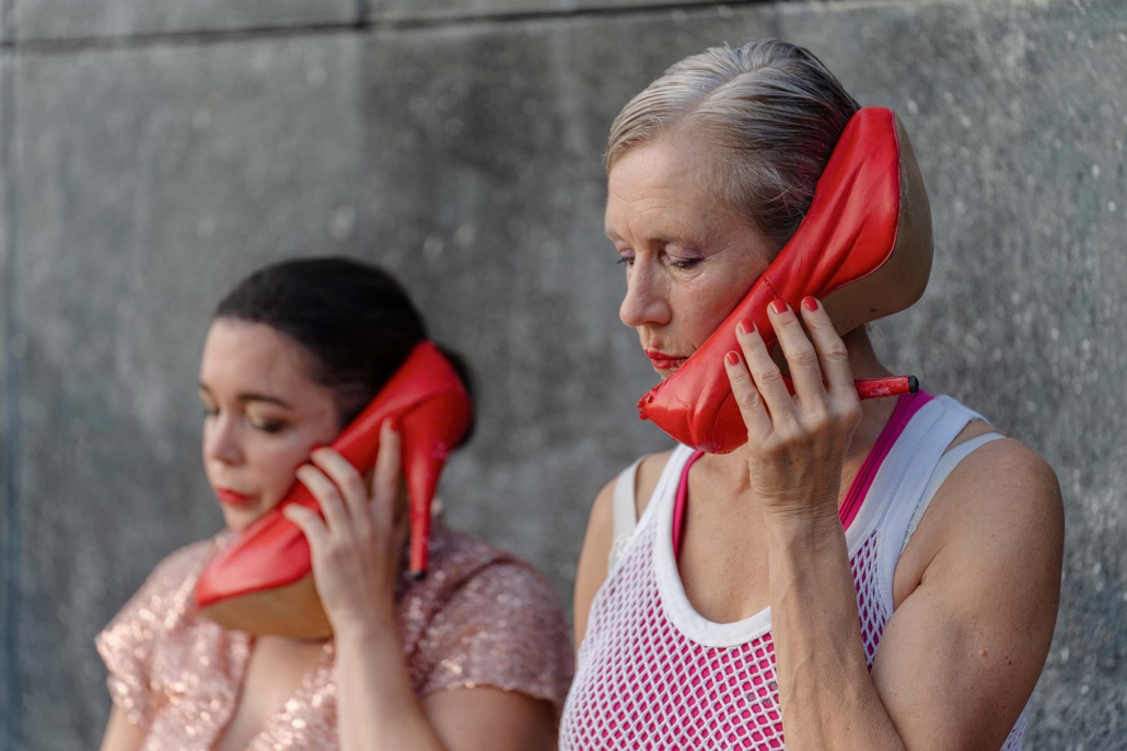 Pilar Murube – Puta Zwei Performerinnen aus „PUTA“ halten rote High-Heels wie Telefonhörer an ihre Ohren.